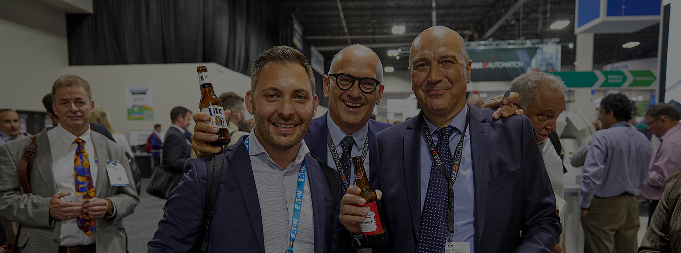 A group of men in formal attire, each holding a beer, engaged in conversation at The Battery Show expo hall
