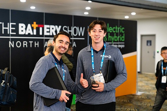  Two men stand in front of a sign reading "The Battery Show," smiling and posing for the camera